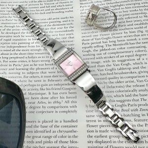 Könnte beinhalten: Eine silberne Guess-Uhr mit einem rechteckigen, rosa Zifferblatt, verziert mit glitzernden Kristallen um die Lünette. Die Uhr hat ein silbernes Armband und wird auf einer bedruckten Seite gezeigt.