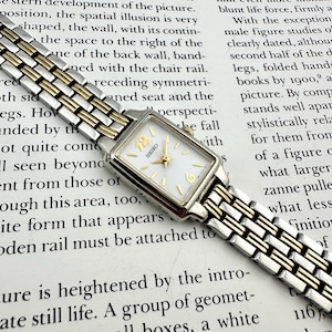 May include: A rectangular Seiko wristwatch featuring a white dial with gold-tone hour markers and hands, including the numerals 12 and 6. The watch has a two-tone metal link bracelet combining silver and gold-tone finishes with a secure clasp.