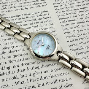 Può includere: Orologio Lorus argentato con quadrante azzurro chiaro iridescente e dettagli di diamanti. L'orologio ha un cinturino argentato e un movimento al quarzo. L'orologio è mostrato su una pagina stampata.