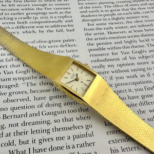 Puede incluir: Un reloj de pulsera Citizen Quartz en tono dorado con caja rectangular y brazalete de malla texturizada integrado. La esfera es de color blanco roto con finos marcadores de hora negros, manecillas negras y el texto Citizen Quartz.