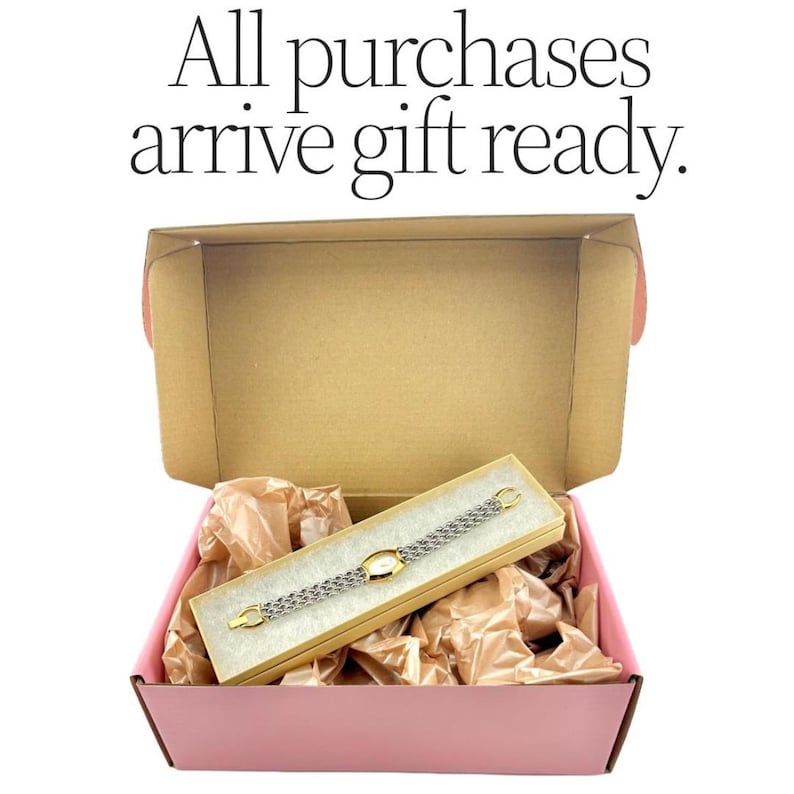 K&ouml;nnte beinhalten: Ein gold- und silberfarbenes Armband in einer Geschenkbox, eingebettet in braunes Packpapier, in einer rosa Box. Der Text "All purchases arrive gift ready" befindet sich oben im Bild.