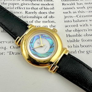 May include: A gold quartz watch with a black leather strap. The watch face has a mother-of-pearl design, gold hands, and a small logo. A classic timepiece.