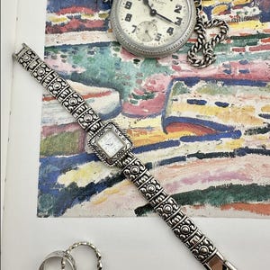 Könnte beinhalten: Eine antike Taschenuhr mit einer Silberkette und eine quadratische Armbanduhr aus Silber werden auf einem farbenfrohen abstrakten Gemälde präsentiert. Zwei Silberringe sind ebenfalls sichtbar. Die Taschenuhr hat ein weißes Zifferblatt mit schwarzen Ziffern.
