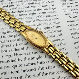 Può includere: Un orologio da polso Pulsar Quartz color oro con quadrante ovale e cinturino a maglie. L'orologio presenta numeri romani come indicatori delle ore e lancette sottili. L'orologio è posto su uno sfondo di testo stampato.