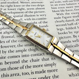 May include: A rectangular Seiko watch with a white face and gold-tone hands and bezel. The watch has a silver and gold-tone band. The watch is on a printed paper background.
