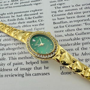 May include: Gold-toned wristwatch with an oval green face, surrounded by a ring of clear crystals. The watch band features a textured design. The watch is set against a backdrop of printed text.