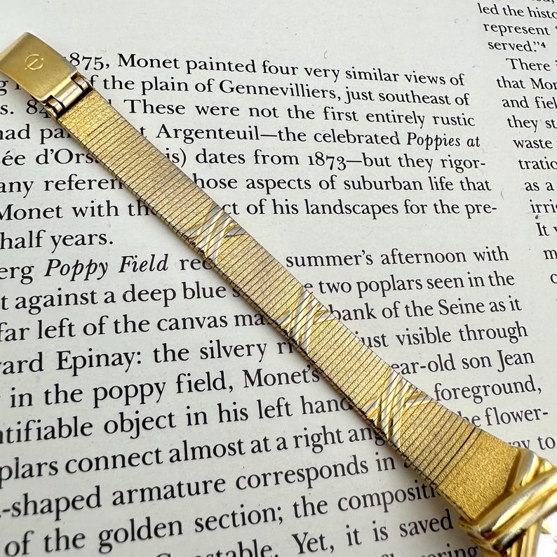 K&ouml;nnte beinhalten: Ein goldfarbenes Uhrenarmband mit einem strukturierten, gewebten Design. Das Armband ist vor einem Hintergrund aus gedrucktem Text platziert. Der Verschluss hat ein kleines, graviertes Emblem. Ein klassisches Accessoire.