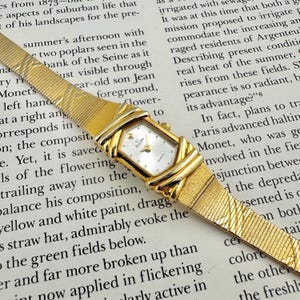 K&ouml;nnte beinhalten: Eine goldfarbene Elgin-Quarzuhr mit rechteckigem Zifferblatt und strukturiertem Armband. Die Uhr hat ein wei&szlig;es Zifferblatt mit goldenen Zeigern und Markierungen. Das Armband hat ein gewebtes Design. Die Uhr befindet sich auf einem Hintergrund mit gedrucktem Text.