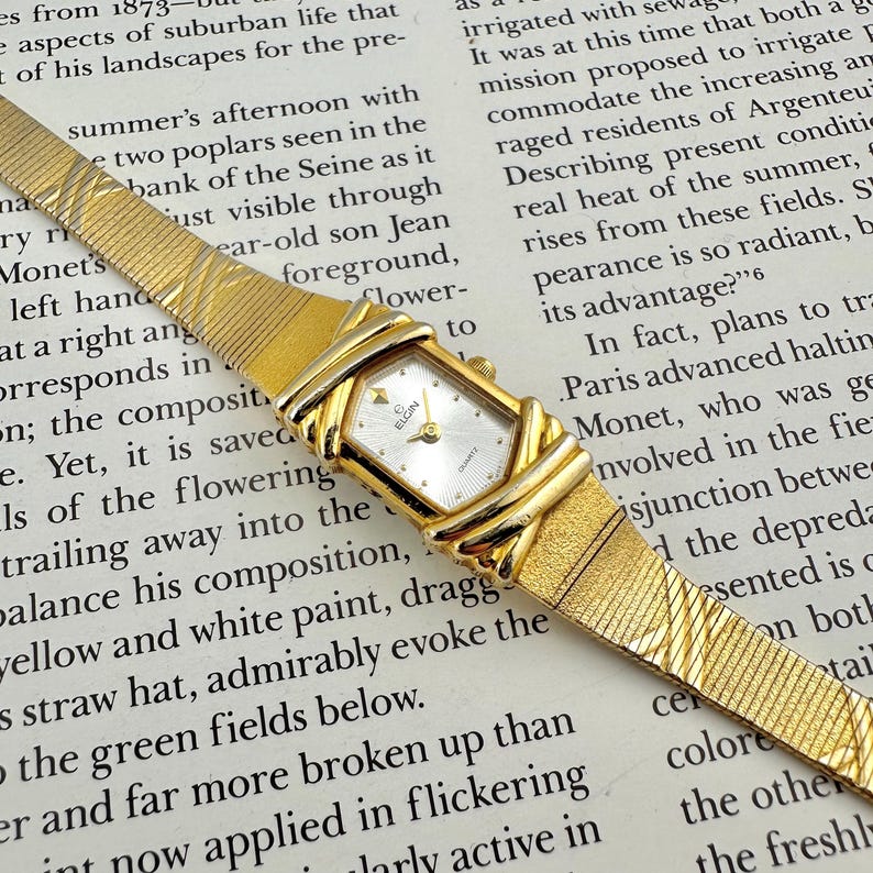 K&ouml;nnte beinhalten: Eine goldfarbene Elgin-Quarzuhr mit rechteckigem Zifferblatt und strukturiertem Armband. Die Uhr hat ein wei&szlig;es Zifferblatt mit goldenen Zeigern und Markierungen. Das Armband hat ein gewebtes Design. Die Uhr befindet sich auf einem Hintergrund mit gedrucktem Text.