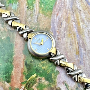 Puede incluir: Un reloj de pulsera Bulova con esfera azul claro y manecillas y detalles dorados. El reloj tiene una correa de eslabones plateada y dorada. La esfera del reloj tiene el logotipo de Bulova.