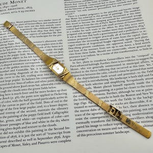 K&ouml;nnte beinhalten: Eine goldene Armbanduhr mit rechteckigem Zifferblatt und Mesh-Armband liegt auf einer bedruckten Seite. Die Uhr hat ein klassisches Design mit einem Goldgeh&auml;use und einem schlichten Zifferblatt. Das Armband ist fein gewebt.
