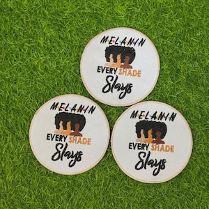 May include: Three circular white fabric patches featuring embroidered text that reads MELANIN EVERY SHADE Slays. The design includes a graphic of three stylized silhouettes with afro hairstyles in black, tan, and brown colours above the text.