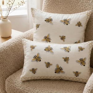 May include: Two decorative pillows with a bee theme. The pillows are off-white with embroidered yellow and black bees. The pillows have a honeycomb border. The pillows are on a beige armchair.