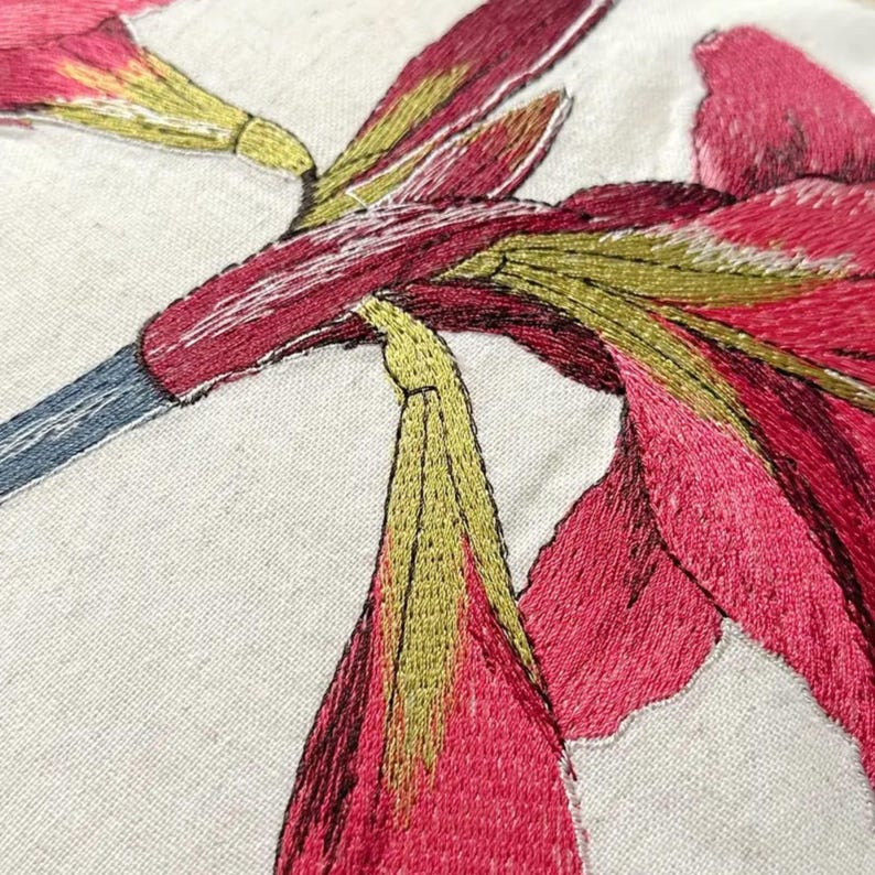 May include: Close-up of an embroidered floral design featuring vibrant pink and red flowers with yellow-green accents on a cream-colored fabric. The embroidery showcases detailed stitching, creating a textured and artistic appearance.