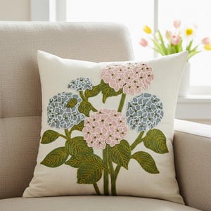 Puede incluir: Cojín cuadrado color crema con un diseño floral bordado. El diseño incluye flores de hortensia rosas y azules con hojas y tallos verdes. El cojín está colocado sobre un sillón beige.