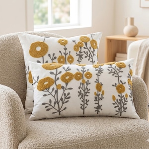 Peut inclure: Deux coussins blancs avec des fleurs jaunes brodées et des tiges grises. L'un est rectangulaire, l'autre carré. Les coussins sont sur un fauteuil beige, créant une décoration intérieure sur le thème floral.