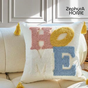 Peut inclure: Un coussin décoratif carré blanc cassé avec le mot "HOME" brodé en rose, jaune, bleu et blanc. Le coussin a des glands jaunes et la marque "ZephyrA HOME" est imprimée dans le coin supérieur droit.