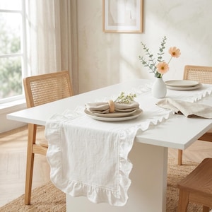 May include: A white rectangular table is set for a meal with a white ruffled table runner. Beige plates, napkins, and a wooden napkin ring are on the table. A vase with flowers and two woven chairs complete the dining scene.
