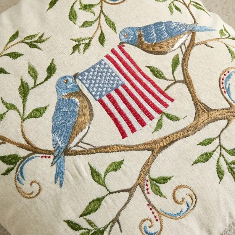 May include: Embroidered pillow featuring two bluebirds perched on a branch, holding an American flag. The design includes green leaves and brown branches against a cream-colored background. The embroidery is detailed and colorful.