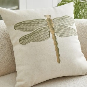 May include: Square decorative pillow with a large embroidered dragonfly design. The dragonfly features olive green wings and a tan body, set against a cream-colored background. The pillow is a square shape and appears to be made of a textured fabric.
