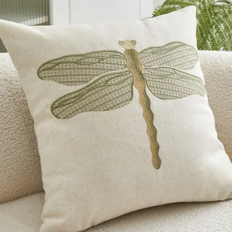 May include: Square decorative pillow with a large embroidered dragonfly design. The dragonfly features olive green wings and a tan body, set against a cream-colored background. The pillow is a square shape and appears to be made of a textured fabric.