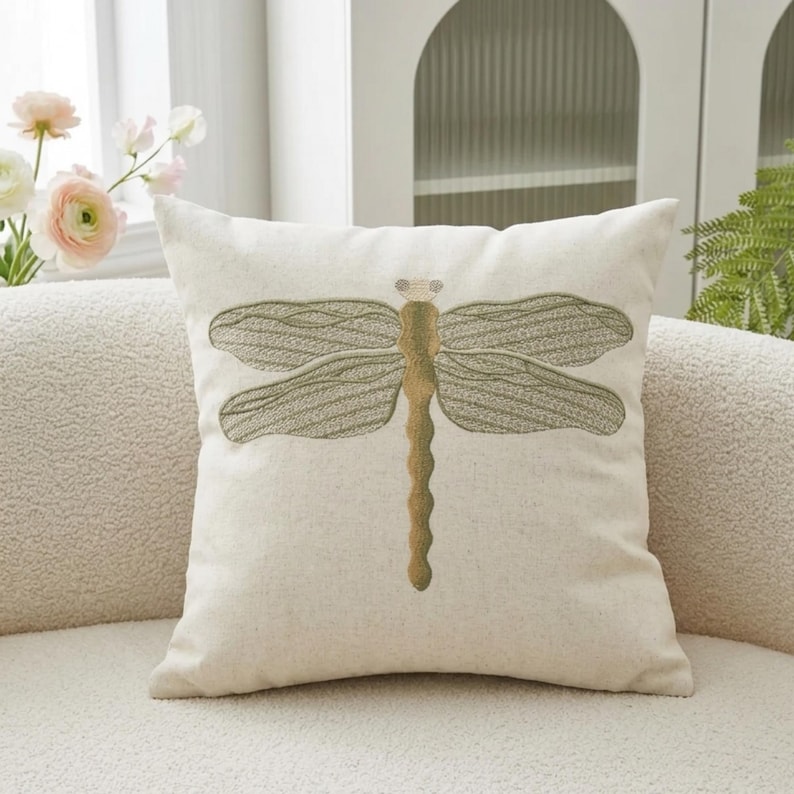 May include: Off-white square throw pillow featuring an embroidered dragonfly design. The dragonfly has olive green wings and a golden-brown body. The pillow is made of a textured fabric and is placed on a white sofa.