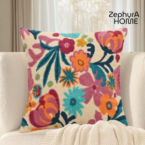 Peut inclure: Coussin carré avec un fond crème et un motif floral coloré. Les fleurs brodées sont dans des tons d'orange, de rose, de turquoise et de jaune. Le coussin est posé sur un canapé blanc avec le texte "ZephyrA HOME" en haut à droite.