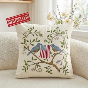 May include: Cream-colored square throw pillow with embroidered design. Two blue birds perch on a brown branch, holding a small American flag. Green leaves and vines surround the birds. The word "BESTSELLER" is in a red rectangle.