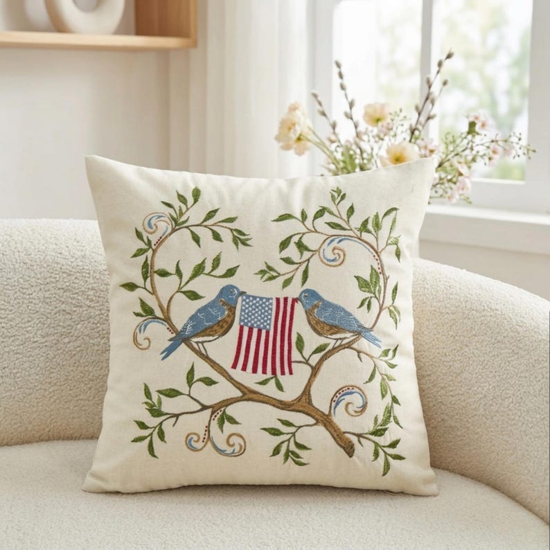 May include: Cream-colored square throw pillow with an embroidered design. Two blue birds perch on a brown branch, holding a small American flag. The branch is surrounded by green leaves and decorative swirls.