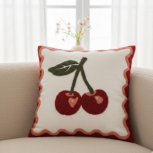 Puede incluir: Cojín cuadrado decorativo con diseño de cerezas sobre fondo blanco. Las cerezas son de color rojo oscuro con una hoja y tallo verdes. El cojín tiene un borde rojo festoneado.