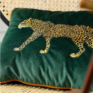 May include: A square, dark green velvet cushion featuring a gold embroidered leopard motif. The leopard is depicted in a walking pose. The cushion is trimmed with a gold border.