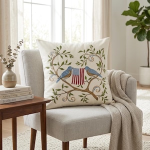 May include: Cream-colored decorative pillow with an embroidered design featuring two blue birds perched on a branch, holding a small American flag. The birds are surrounded by green leaves and vines. The pillow is placed on a gray chair.