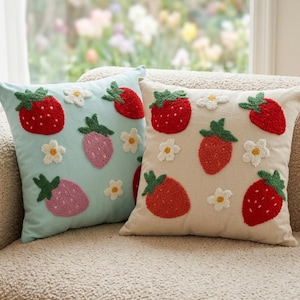 Peut inclure: Deux coussins décoratifs avec des motifs brodés de fraises et de fleurs. L'un est bleu clair, l'autre beige. Les fraises sont rouges et roses avec des feuilles vertes. Des fleurs blanches sont éparpillées.