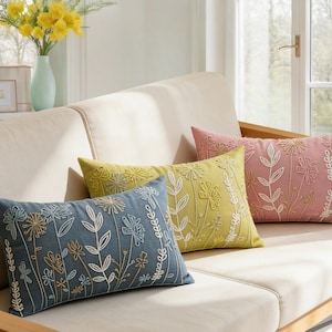 Op de afbeelding: Vier geborduurde sierkussens in blauw, geel en roze, geplaatst op een beige bank. Elk kussen heeft bloempatronen. Een vaas met gele bloemen is zichtbaar, wat de inrichting van de kamer verbetert.