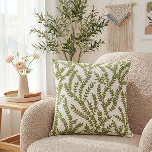 Puede incluir: Un cojín decorativo cuadrado con fondo blanco y un patrón de hojas bordadas en verde. El cojín está colocado sobre un sillón beige. Un jarrón con flores y una planta decorativa están en el fondo.