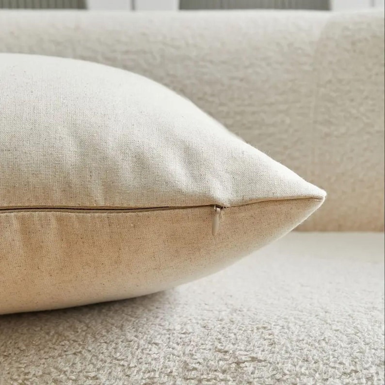 May include: Close-up of a beige pillow with a zipper closure. The pillow is made of a textured fabric and is resting on a matching beige surface. The pillow's corner is in focus, showing the fabric's weave.