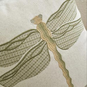 May include: Close-up of a decorative pillow with an embroidered dragonfly design. The dragonfly is rendered in shades of green and tan, with detailed wing patterns and a textured body. The pillow has a neutral, off-white background.