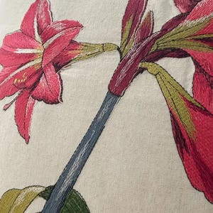 May include: Close-up of an embroidered floral design featuring a vibrant pink amaryllis flower. The embroidery showcases detailed stitching in shades of pink, green, and blue on a neutral fabric background. The design highlights the flower's petals and stem.