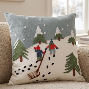 May include: Embroidered square pillow with a winter scene. The design features two figures pulling a sled up a snowy hill, surrounded by green pine trees and falling snowflakes. The pillow has a light blue background.