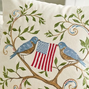May include: Embroidered pillow featuring two bluebirds perched on a branch, holding an American flag. The design includes green leaves and decorative swirls, all on a cream-colored background. The pillow has a patriotic theme.