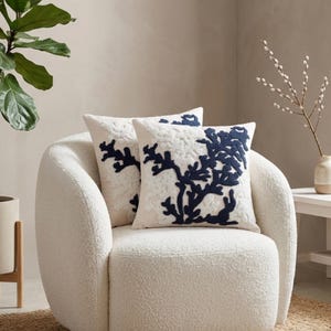 May include: Two square cushions with a cream background and a navy blue coral design. They are placed on a white, textured armchair. The room has a neutral colour scheme with a plant and vase in the background.