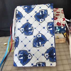 Puede incluir: Una cubierta de libro con un patrón repetido de escudos y estrellas azules y blancos de Ravenclaw. La cubierta está atada con cintas de colores. Una pila de libros con cubiertas florales y estampadas es visible en el fondo.