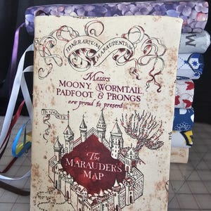 May include: A stack of fabric-covered journals, with the top one featuring a detailed illustration of The Marauder's Map from Harry Potter. The map is in shades of brown and red, with text and a castle design. Ribbons of various colors are attached.