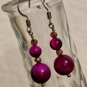May include: A pair of dangle earrings featuring large, round, deep magenta beads. The earrings have a bronze-colored metal hook and wire design, with smaller, faceted beads in between the larger ones. The earrings are displayed against a blurred background.