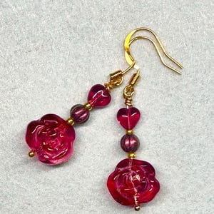 May include: A pair of dangle earrings featuring gold-colored hooks and wire. Each earring has a red rose-shaped bead, a heart-shaped bead, and several smaller beads in shades of red and gold. The earrings are displayed against a light gray background.