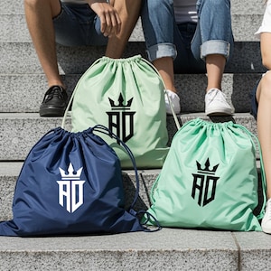 May include: Three drawstring bags in navy blue, light green, and mint green. Each bag features a white or black logo with a crown design and the letters "AD". The bags are sitting on concrete steps.