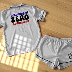 May include: A matching pajama set in a light silver satin fabric. The top has the text "Dreaming of ZERO Deductions" in blue, black, and red. The shorts say "cheer COACH" in black.