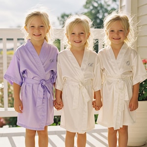 May include: Three young children wearing matching satin robes. One robe is lavender, and the other two are white. Each robe has a matching tie and a single initial embroidered on the front. The children are smiling and holding hands.
