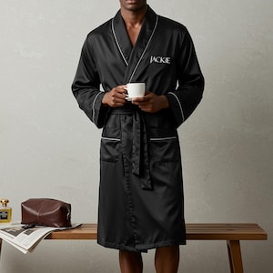 May include: A black satin robe featuring white piping along the shawl collar, cuffs, and pockets. The word JACKIE is embroidered in white thread on the left chest. The robe includes a matching fabric tie belt at the waist and long sleeves.