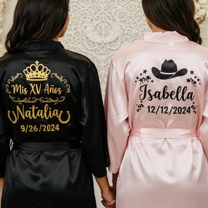 May include: Two satin robes, one black and one pink, with personalized gold text. The black robe has a crown design and reads "Mis XV Años Natalia 9/26/2024." The pink robe features a cowboy hat and reads "Isabella 12/12/2024."